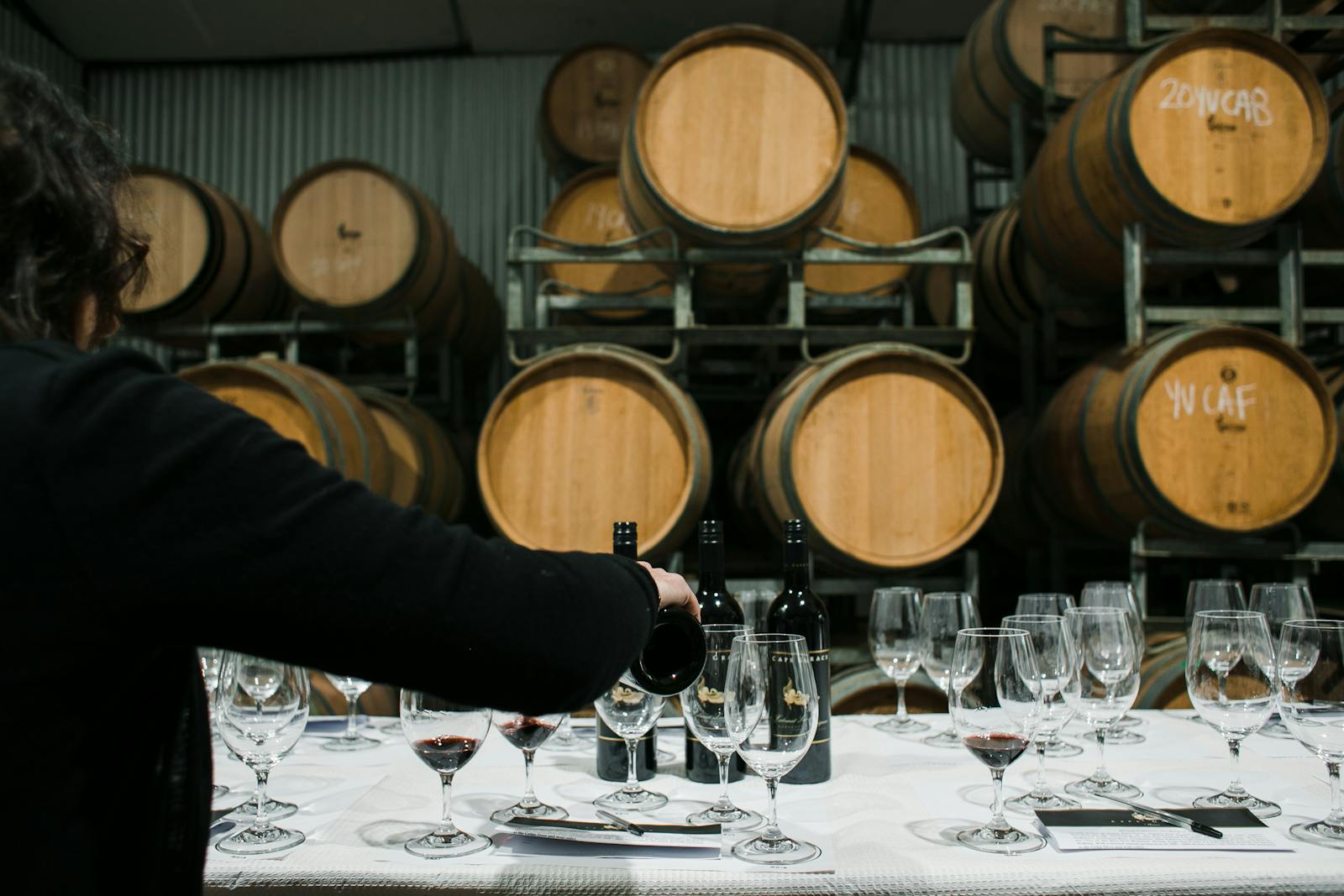 Back view of anonymous sommelier pouring wine from bottle into wineglass while standing at table with glassware in winery against barrels